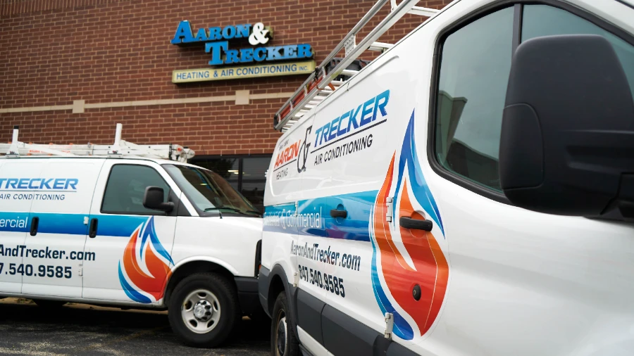 Two Aaron & Trecker Heating & Air Conditioning vans are parked outside a brick building with the company’s logo and contact information visible on the vehicles.
