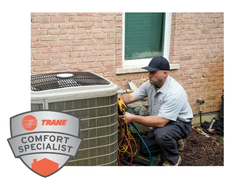 An HVAC technician is checking an outdoor AC unit with gauges next to a brick house. A “Trane Comfort Specialist” badge is shown in the bottom left corner.