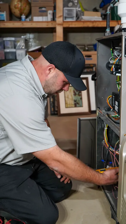 A technician in a gray shirt and black cap repairs wiring inside an open furnace unit in a basement.