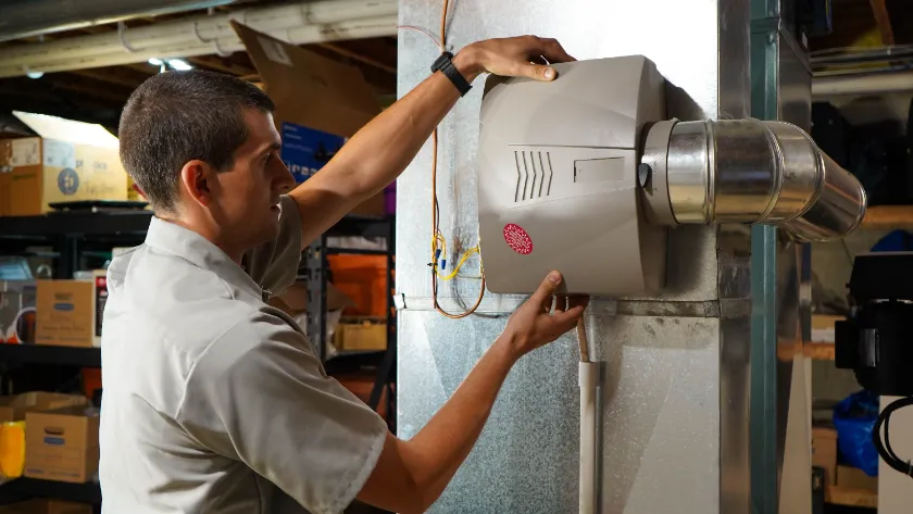 A man installs or repairs a humidifier unit attached to a metal air duct in a basement with shelves and storage boxes in the background.