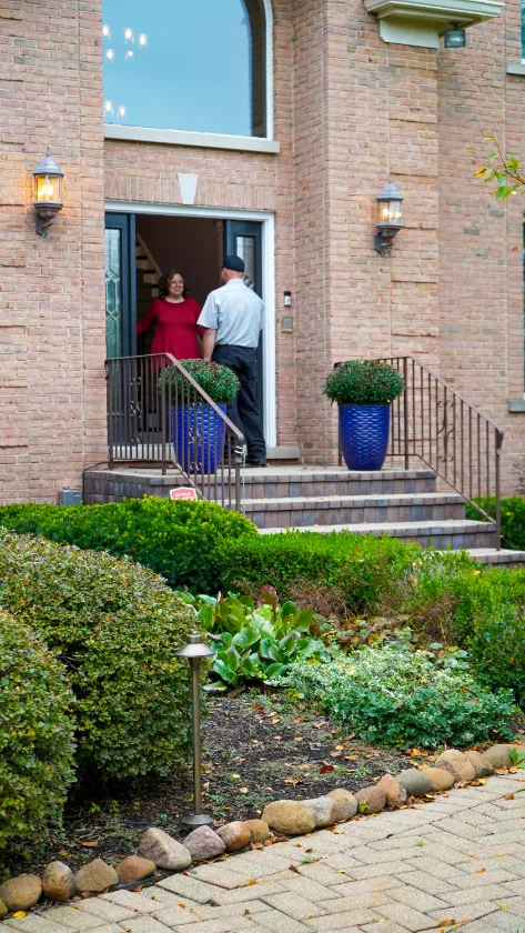 A man stands at the front door speaking with a woman inside a brick house, surrounded by bushes, potted plants, and a stone-edged walkway.