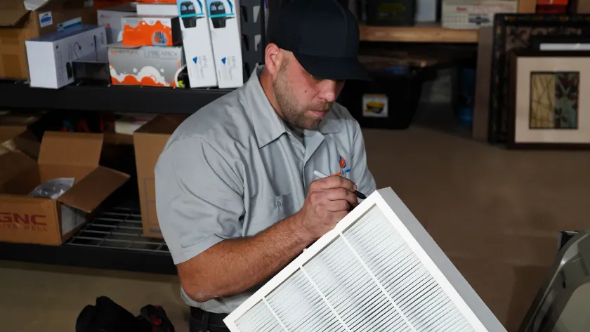 A man in a gray uniform and cap is working on a white air filter in a workshop with shelves, boxes, and tools in the background.
